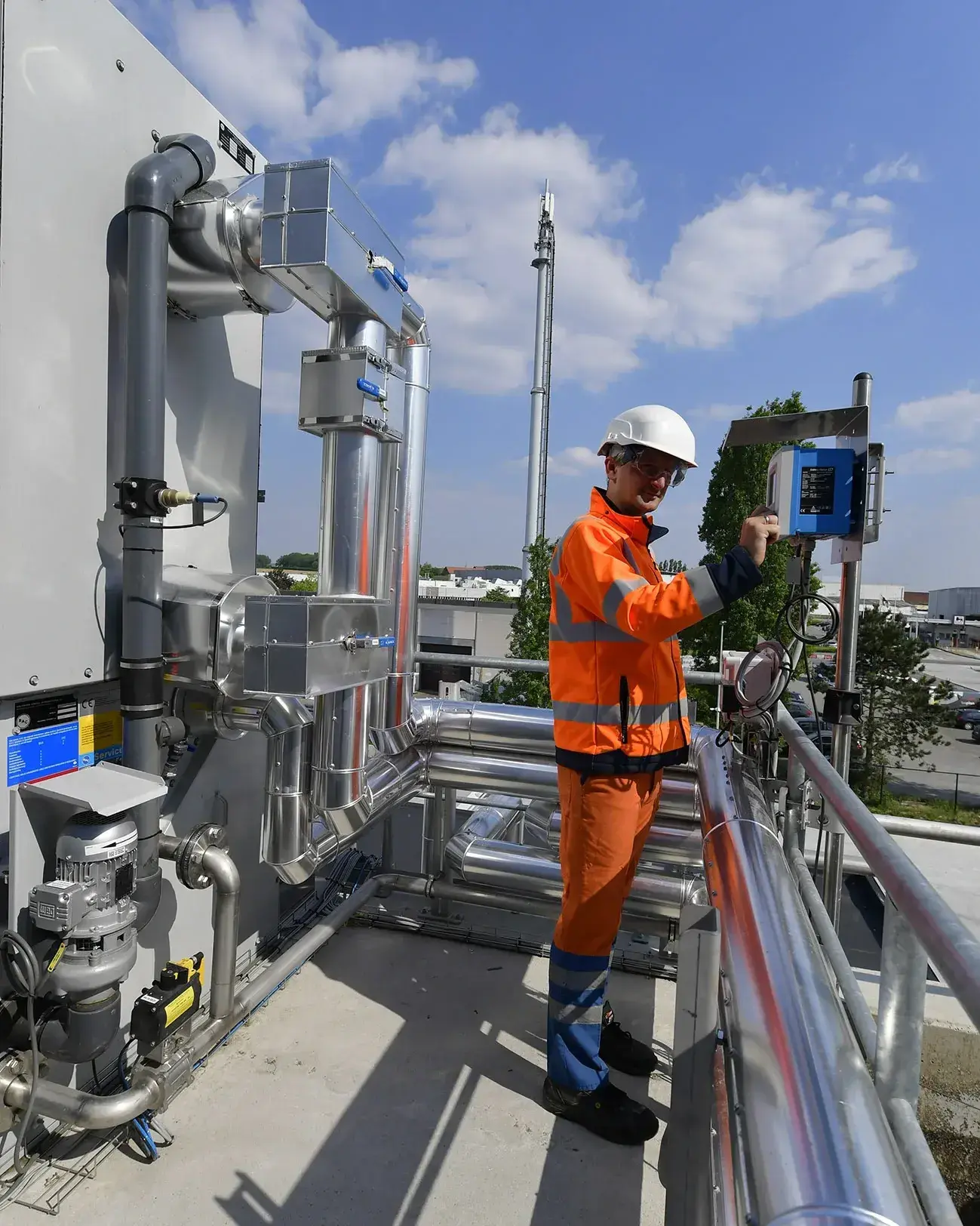 Technicus in een oranje veiligheidsoutfit en witte helm bedient een controlepaneel op een industriële installatie. Hij staat op een verhoogd platform omgeven door roestvrijstalen leidingen en ventilatiesystemen. De achtergrond toont een industrieterrein met gebouwen en een blauwe lucht met enkele wolken.