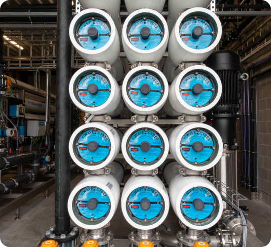 Een industriële waterfiltratie-installatie met twaalf grote, horizontale filtratie-units met blauwe eindkappen, gerangschikt in een symmetrisch patroon. De installatie is omgeven door leidingen, kleppen en een pomp, wat wijst op een geavanceerd waterzuiveringssysteem. De achtergrond toont extra buizen en technische structuren binnen de fabriekshal.