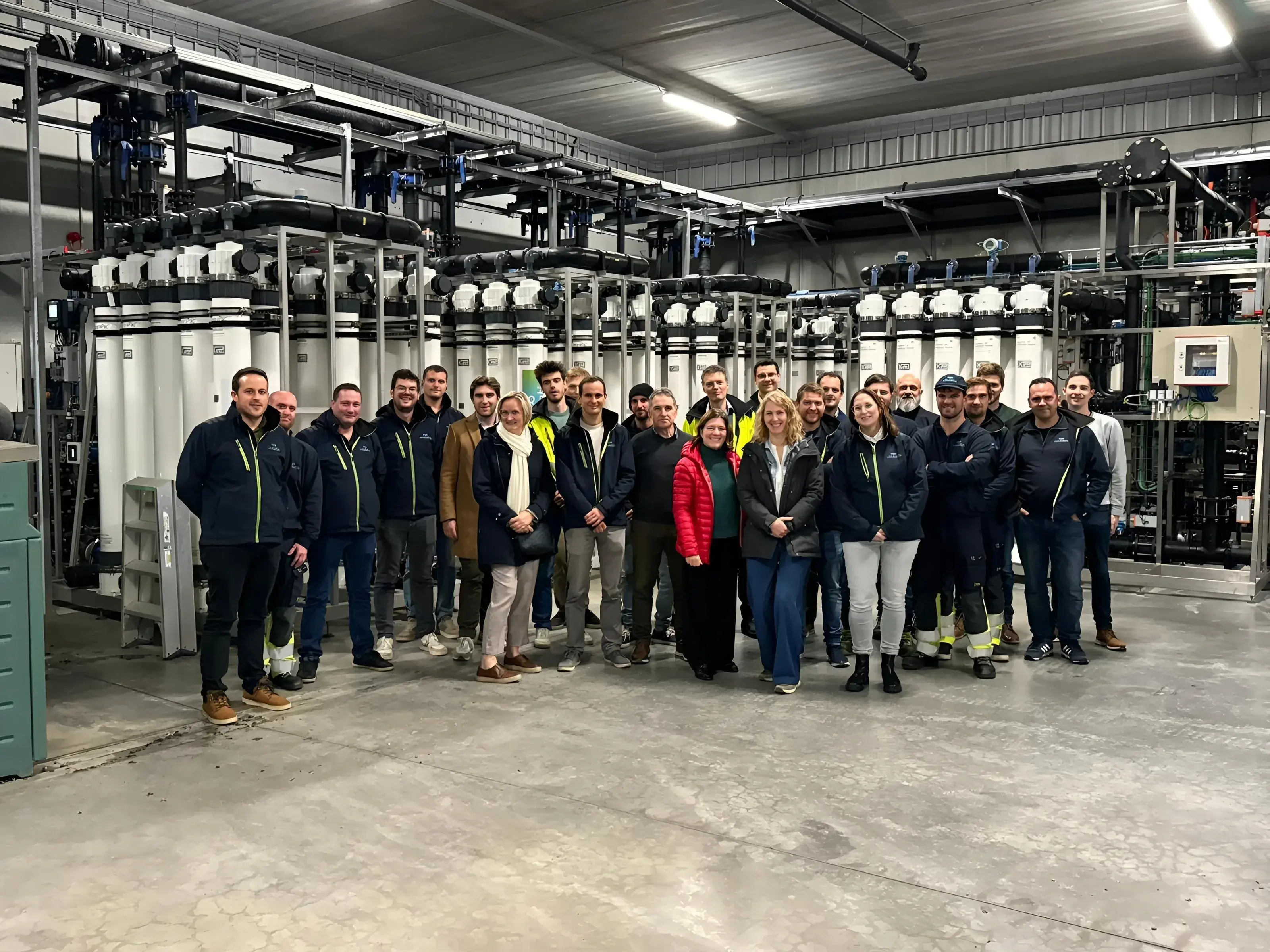 Een groep van ongeveer 20 medewerkers poseert samen in een industriële omgeving, omringd door grote waterfiltratie-installaties met buizen en technische apparatuur. Ze dragen een mix van werk- en casual kleding, waaronder veiligheidsjassen en bedrijfskleding. De sfeer is informeel en collegiaal, met een goed verlichte werkruimte en een betonnen vloer.