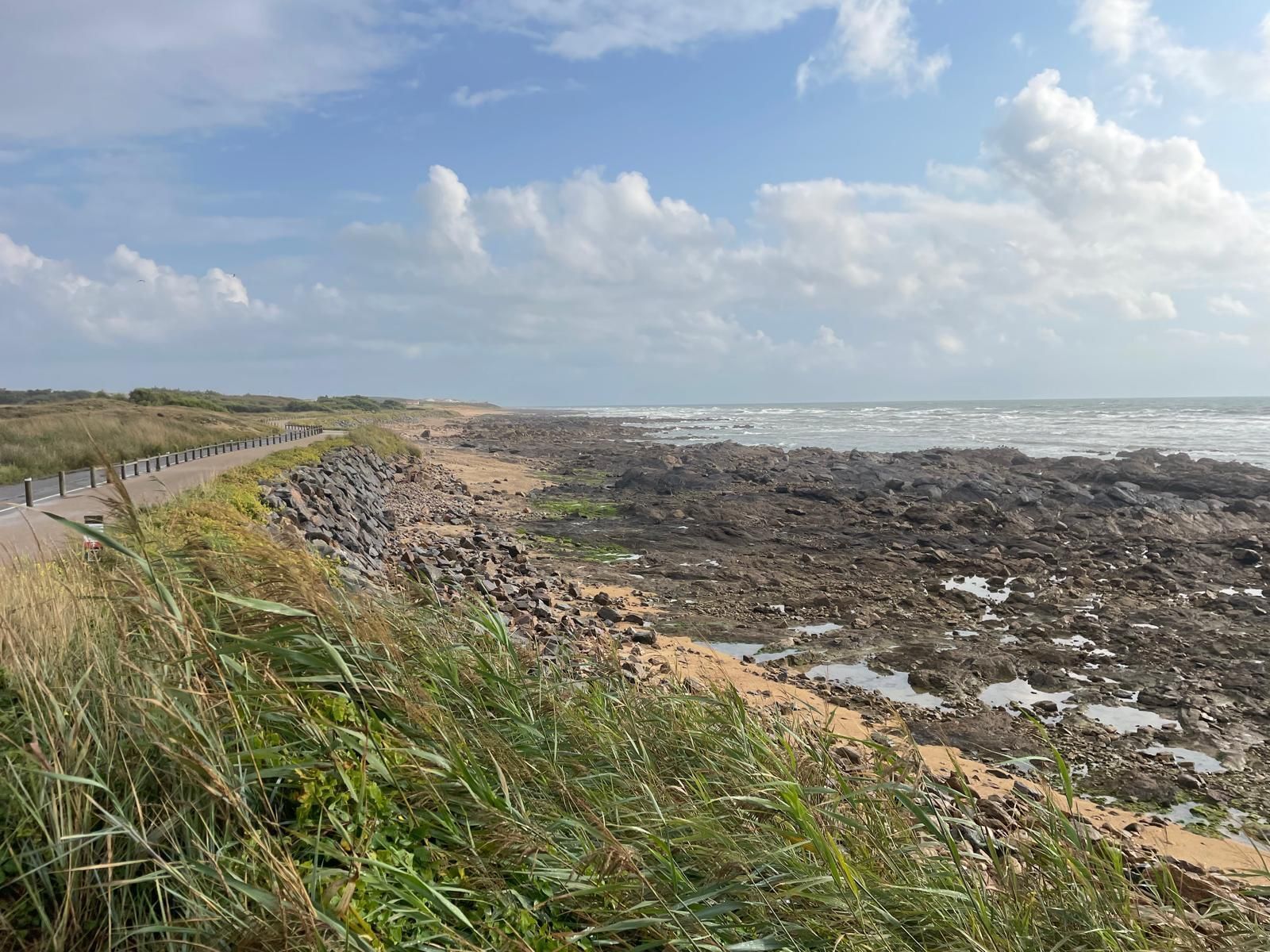 Kustlandschap met een ruige, rotsachtige kustlijn en een smalle strook zandstrand. Op de voorgrond wuiven groene grassen in de wind langs een fietspad dat parallel aan de kust loopt. De zee heeft kleine golven en de lucht is deels bewolkt met blauwe stukken zichtbaar tussen de wolken.