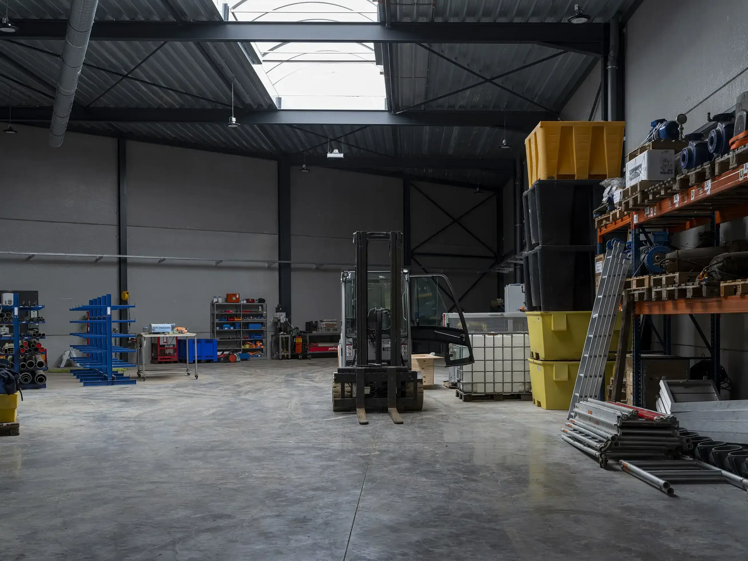 Ruime industriële loods met een betonnen vloer en een hoog plafond met metalen spanten. In het midden staat een heftruck, omringd door stellingen met buizen, containers en gereedschappen. Links zijn blauwe rekken met materialen en een werkbank zichtbaar. Een dakraam laat natuurlijk licht binnen, terwijl de ruimte verder sober verlicht is.