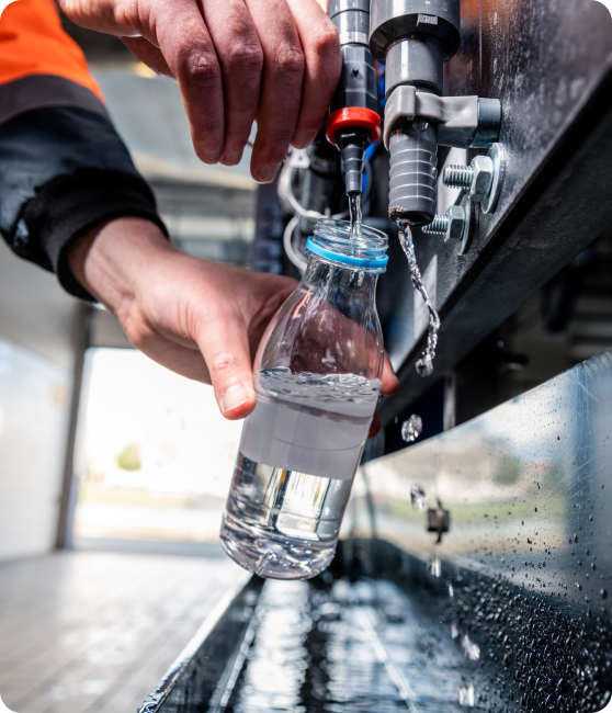 Een persoon in een oranje veiligheidsjas vult een kleine plastic fles met helder water uit een industriële kraan. De hand houdt de fles stevig vast terwijl het water stroomt. De metalen installatie heeft meerdere leidingen en kranen, met waterdruppels zichtbaar op het oppervlak.