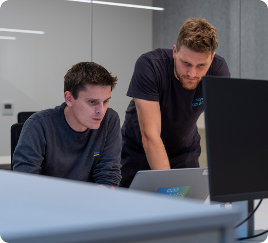 Twee jonge mannen werken samen in een moderne kantooromgeving. De man links zit en kijkt geconcentreerd naar een computerscherm, terwijl de man rechts, gekleed in een azulatis-shirt, over een laptop gebogen staat. De ruimte heeft een minimalistisch ontwerp met glazen wanden en strakke verlichting.