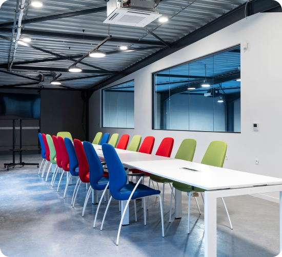 Een moderne vergaderruimte met een lange witte tafel, omringd door kleurrijke stoelen in blauw, rood en groen. De ruimte heeft een industriële uitstraling met een metalen plafond, zichtbare buizen en grote ramen die uitkijken op een andere hal. De betonnen vloer en heldere verlichting geven de kamer een strakke en functionele uitstraling.