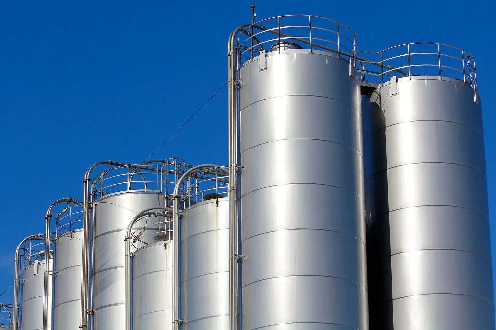 Een rij grote, zilverkleurige opslagsilo's van metaal, glanzend in het zonlicht tegen een heldere blauwe lucht. De silo's hebben relingen en ladders aan de bovenkant, wat wijst op industriële opslag of verwerking, mogelijk voor vloeistoffen of bulkgoederen.