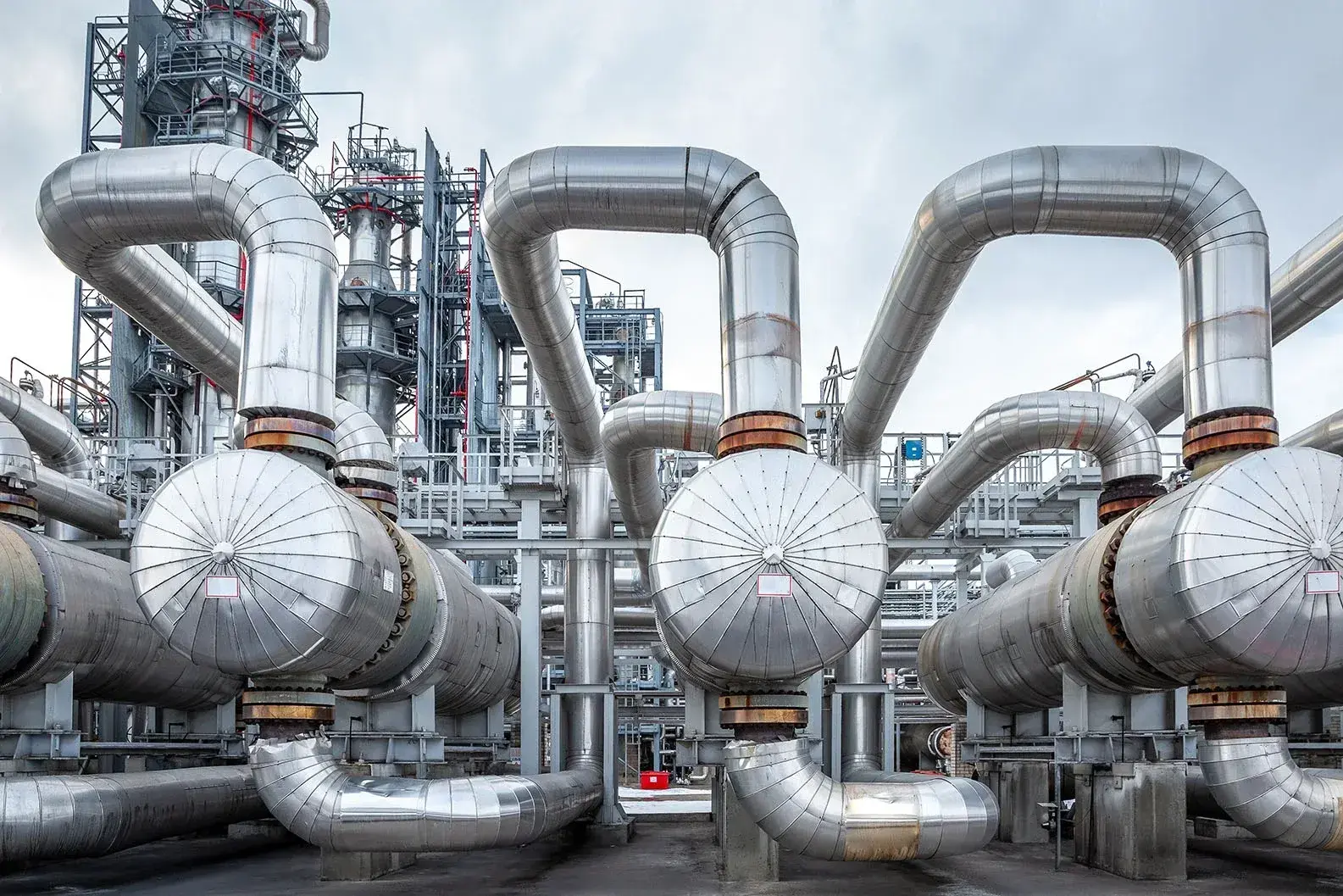 An industrial installation with a complex network of large, silver -colored pipelines that bend in different directions. The metal tubes are equipped with round ends and connected to valves and supporting structures. In the background, extra pipes and steel structures are visible against a cloudy air.