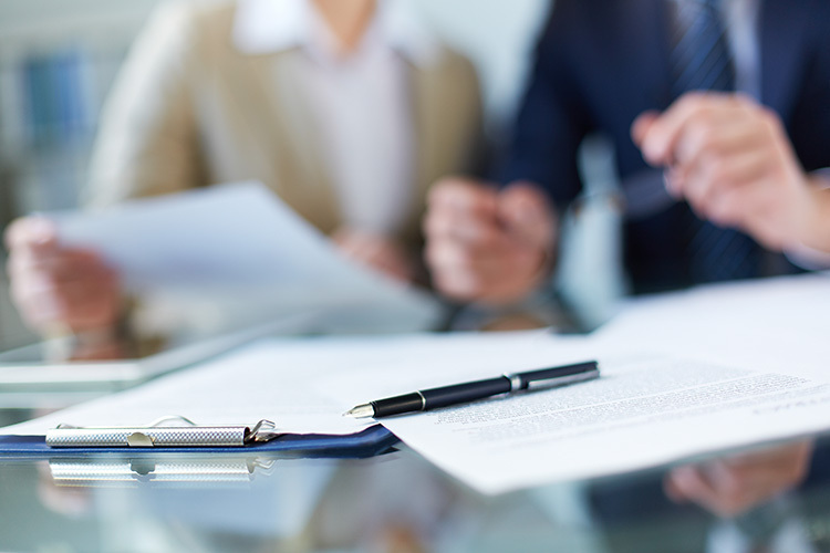 Zakelijke bespreking met twee personen in formele kleding, waarbij documenten worden doorgenomen. Op de voorgrond liggen een pen, een clipbord en een contract op een glazen tafel. De focus ligt op de documenten, terwijl de personen op de achtergrond onscherp zijn.