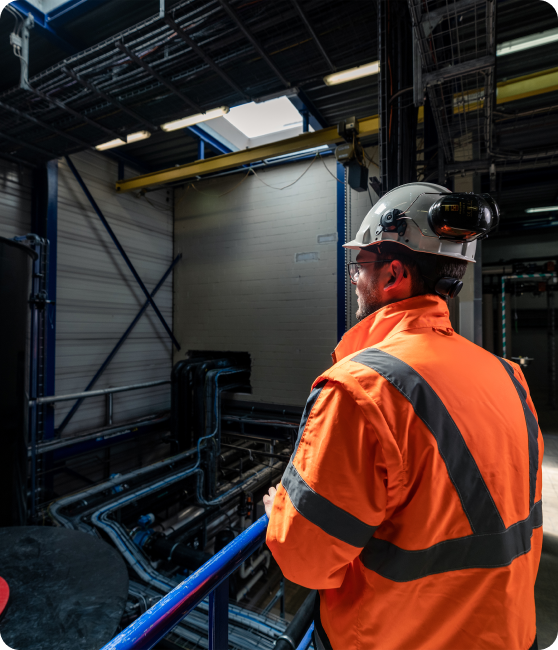 Een man in een oranje veiligheidsjas met reflecterende strepen en een helm met een hoofdlamp kijkt uit over een industriële installatie. Hij staat bij een blauwe railing en observeert een ruimte met metalen buizen, leidingen en een hoge muur met een gesloten poort. Natuurlijk licht valt binnen via een opening in het dak.