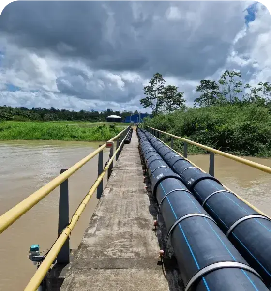 Een betonnen brug met grote zwarte waterleidingen loopt over een rivier, geflankeerd door gele veiligheidsleuningen. De brug leidt naar een kleine structuur in de verte, omringd door groen landschap en bomen. De lucht is deels bewolkt met donkere wolken.