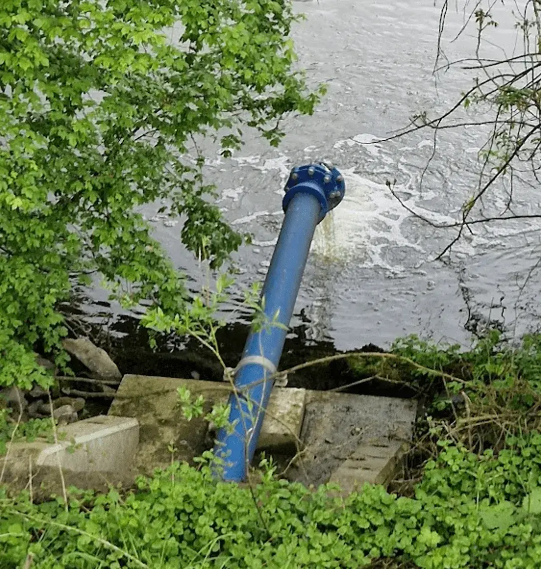 Blauwe afvoerbuis die uitmondt in een water, omringd door groene begroeiing. Het wateroppervlak vertoont lichte stroming en schuimvorming. De buis is bevestigd op betonnen structuren en loopt schuin naar beneden richting het water.