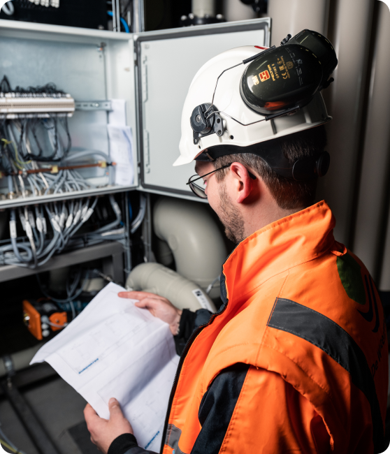 Een technicus in een oranje veiligheidsjas en een witte helm met gehoorbescherming inspecteert een open elektrisch paneel vol bedrading. Hij houdt een document vast met technische schema’s en bestudeert de installatie aandachtig. De omgeving bevat industriële leidingen en apparatuur.