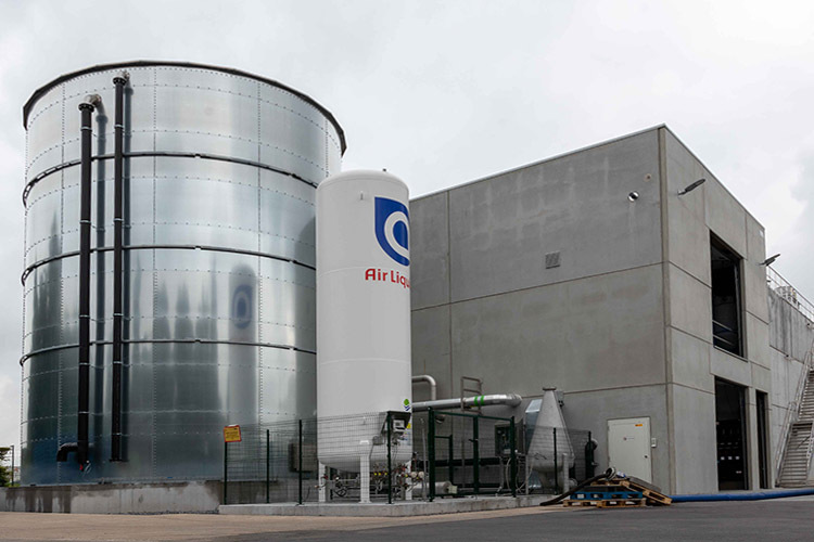 Industriële installatie met een grote metalen opslagtank, een witte gastank met het Air Liquide-logo en een betonnen gebouw. Er zijn zwarte leidingen langs de opslagtank en een omheind gebied met technische apparatuur.