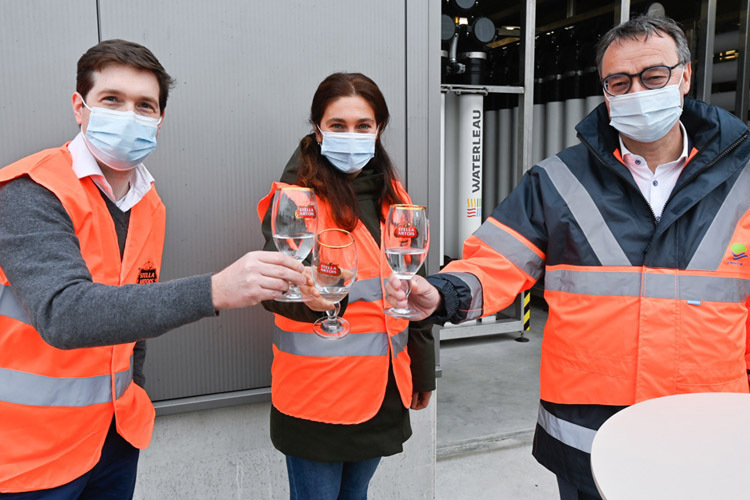 Drie personen in oranje veiligheidsvesten en mondmaskers proosten met glazen water voor een industriële installatie.