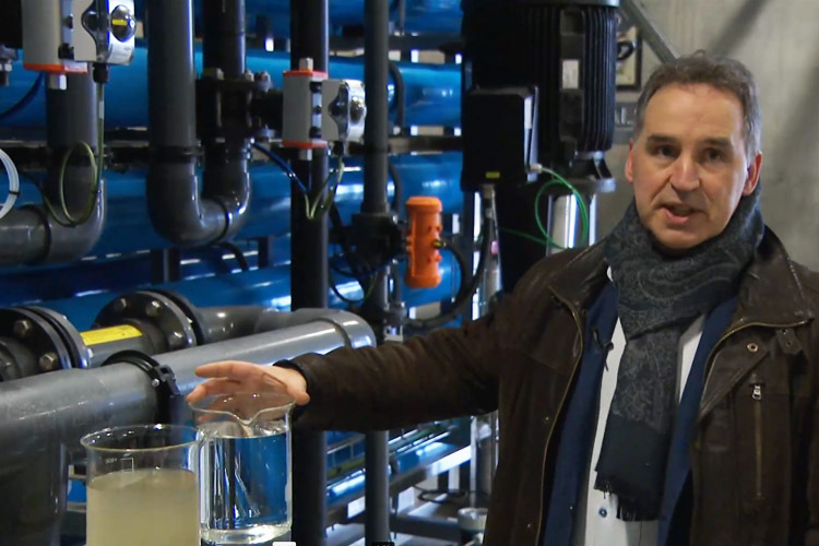 Man geeft uitleg in een industriële waterzuiveringsinstallatie. Hij wijst naar twee glazen cilinders met water, één troebel en één helder, ter demonstratie van het zuiveringsproces. Op de achtergrond zijn blauwe en zwarte leidingen, pompen en regelapparatuur zichtbaar.