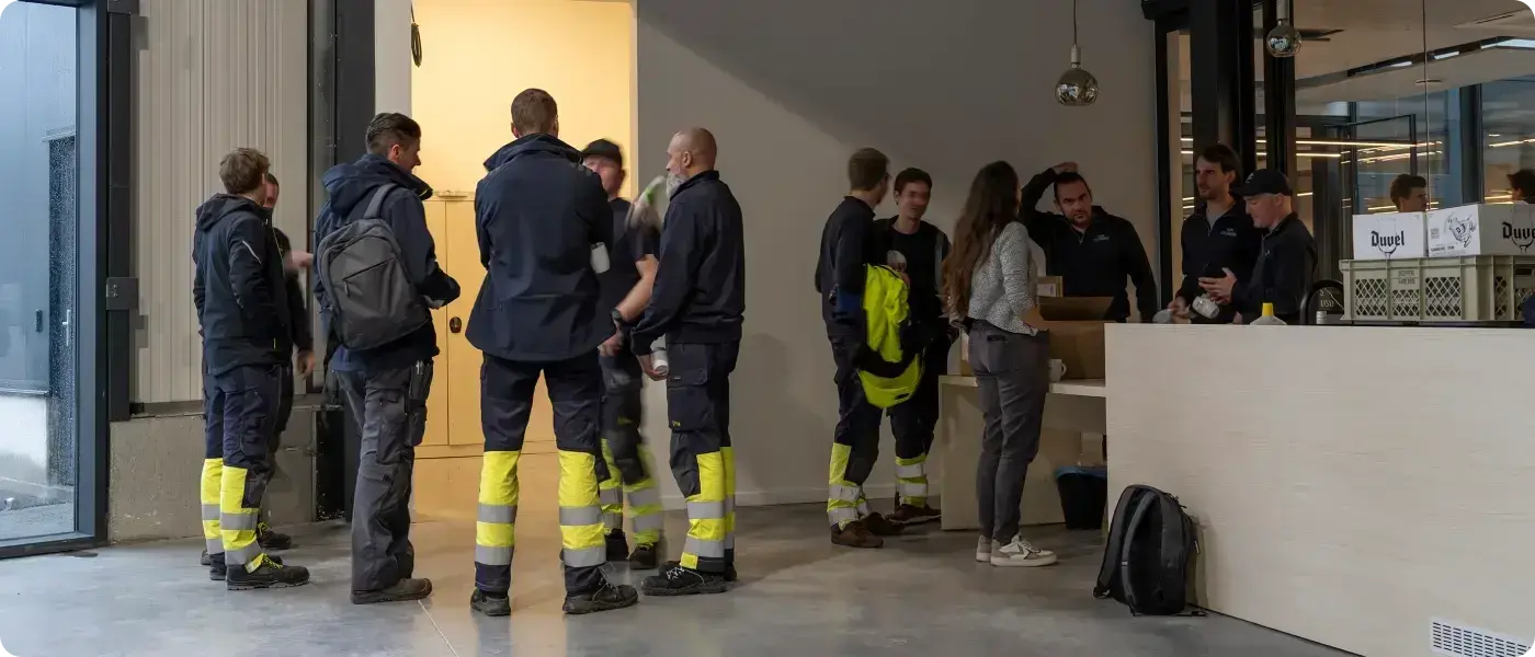 Een groep werknemers in donkerblauwe werkoutfits met reflecterende gele strepen staat samen in een open ruimte, mogelijk een kantine of ontvangstruimte. Sommigen dragen rugzakken, terwijl anderen met collega's in gesprek zijn.