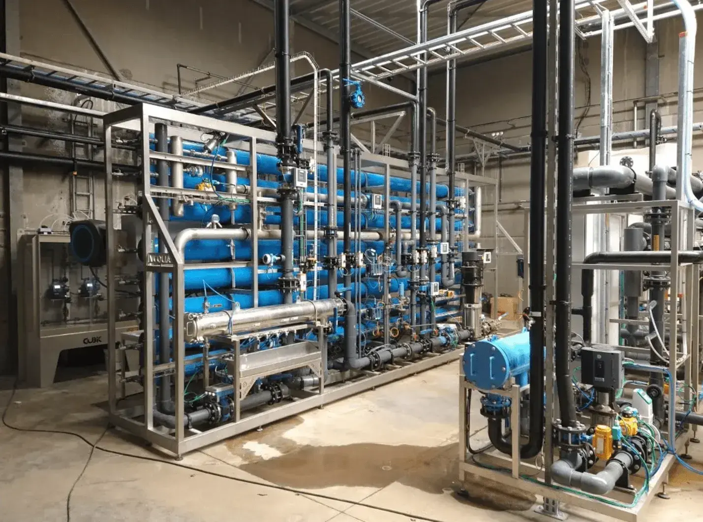 Industriële waterzuiveringsinstallatie met een complex netwerk van blauwe en zwarte leidingen, pompen en kleppen, gemonteerd in roestvrijstalen frames. De installatie bevindt zich in een fabriekshal met betonnen vloeren en metalen structuren. Op de achtergrond zijn extra buizen en technische apparatuur zichtbaar.