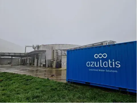 Industriële installatie op een regenachtige dag met een grote, ronde opslagtank en meerdere leidingen op de achtergrond. Op de voorgrond staat een felblauwe container met het Azulatis-logo en de slogan "Unlimited Water Solutions." De natte grond en grijze lucht geven de scène een mistige sfeer.