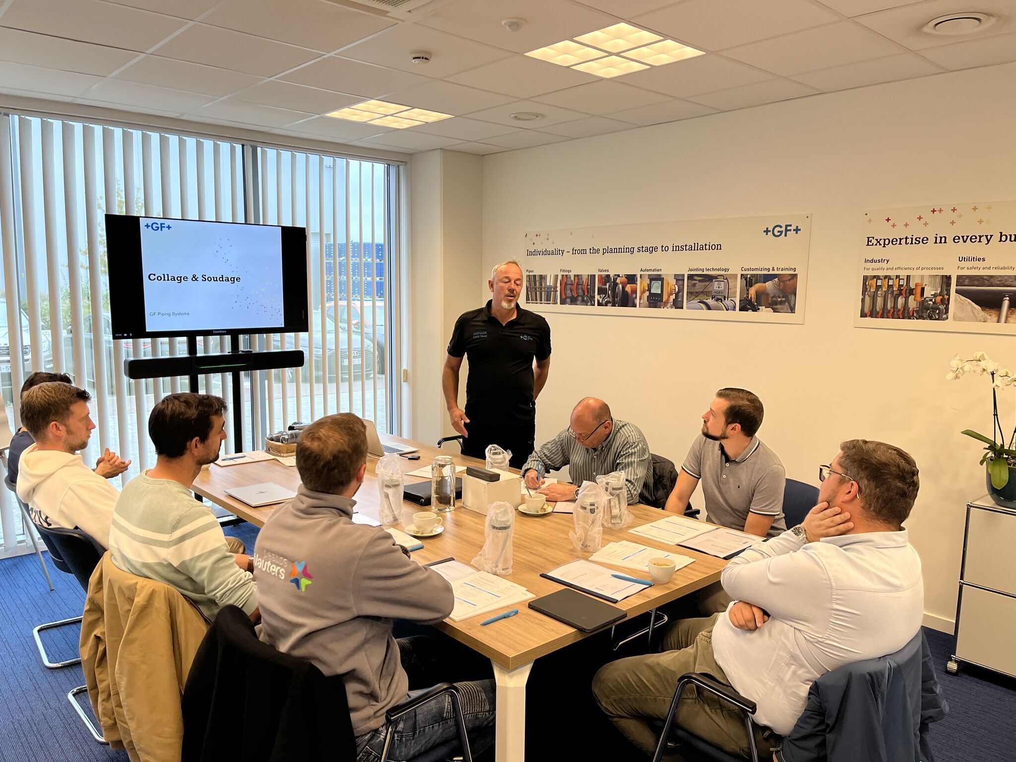 Zakelijke vergaderruimte met zeven deelnemers. Een man in een zwart poloshirt, staat aan het hoofd van de tafel en spreekt de groep toe. De deelnemers, gekleed in casual en zakelijke kleding, zitten rond een houten tafel met notitieblokken, pennen en waterkaraffen. Een groot scherm toont een presentatie met de titel "Collage & Soudage" en het logo van +GF+. De ruimte heeft een moderne uitstraling met blauwe vloerbedekking, grote ramen met verticale jaloezieën en bedrijfsbanners aan de muur met teksten en afbeeldingen over expertise en productieprocessen.