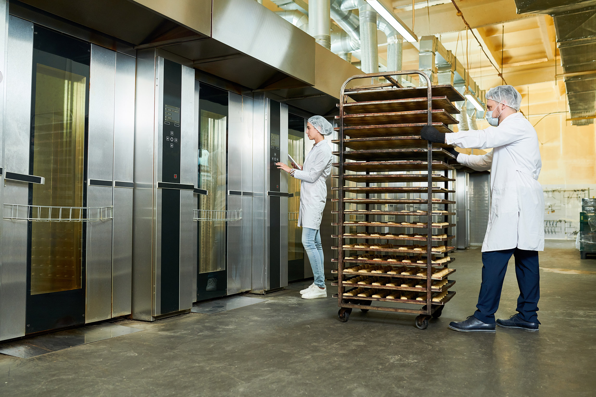 Industriële omgeving met twee werknemers in witte jassen, haarnetjes en handschoenen. Een werknemer bekijkt een grote, roestvrijstalen oven, terwijl de andere een kar met meerdere bakplaten vol producten voortduwt. De ruimte heeft een moderne, schone uitstraling met ventilatiebuizen en metalen ovens op de achtergrond.