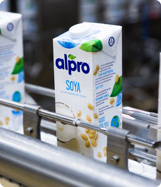 Een pak Alpro Soya drink staat op een productielijn in een fabriek. De verpakking is wit met blauwe en groene accenten en toont sojabonen en een glas sojadrank. Op de achtergrond zijn meer pakken zichtbaar, licht vervaagd, terwijl ze door de metalen transportbaan bewegen.