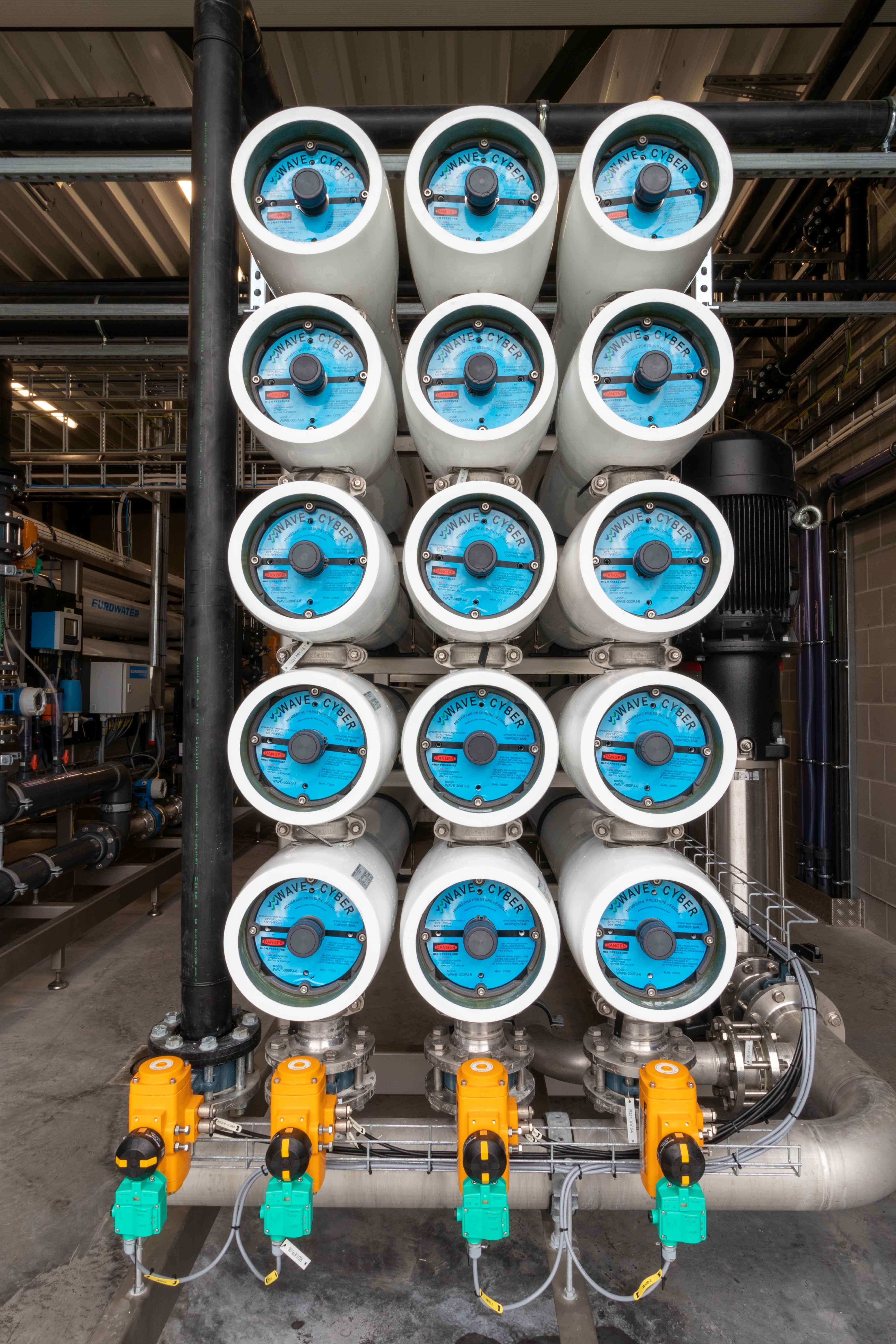 Een industriële waterfiltratie-installatie met 16 horizontale filterunits, elk met een blauwe eindkap en aangesloten op een netwerk van leidingen. Onderaan bevinden zich gele kleppen en regelaars voor de waterstroom. De installatie is gemonteerd in een metalen frame binnen een fabriekshal met buizen en technische apparatuur op de achtergrond.