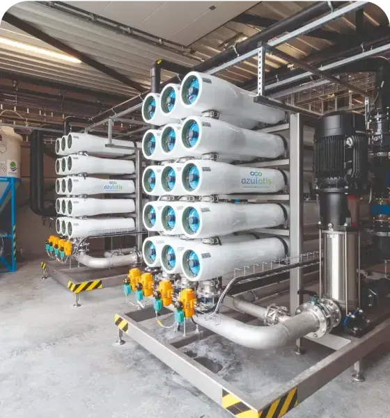 Een industriële waterfiltratie-installatie met meerdere witte filterunits, elk met blauwe eindkappen, gemonteerd op een metalen frame. Gele kleppen en roestvrijstalen leidingen verbinden de onderdelen. De installatie bevindt zich in een fabriekshal met betonnen vloeren en metalen buizen aan het plafond.