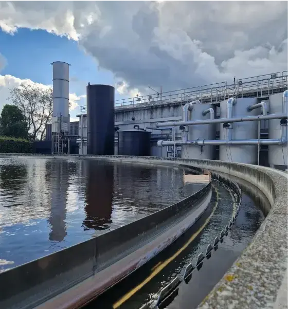 Een waterzuiveringsinstallatie met een groot, rond bezinkingsbassin waarin water wordt gefilterd. Langs de rand loopt een metalen geleider met opvangsysteem. Op de achtergrond staan industriële opslagtanks, leidingen en een fabriek, onder een deels bewolkte lucht.